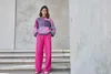 A woman stands on steps in front of a concrete wall, wearing a colorful sweater and pink pants. Produktbild-Vorschau