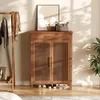 A wooden shoe cabinet with slatted doors and a drawer, placed in an entryway with a coat rack and a plant. Náhľad obrázku produktu