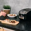 A person is using chopsticks to pick up sushi from a wooden board, with a rice cooker in the background. Produktbild-Vorschau