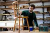 A man is working in a workshop, applying finish to a wooden chair. A blue radio is on the table next to him. Produktbild-Vorschau