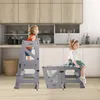 Two children are in a kitchen setting. One child stands on a gray learning tower, looking to the left. Another child sits on a lower gray platform to the right, looking to the right. The kitchen has wooden cabinets and white countertops. Produktbild-Vorschau