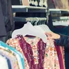 A person's hand is holding a white hanger with a floral patterned dress on it, in a clothing store. Produktbild-Vorschau