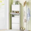 A white shoe cabinet with multiple flip-down compartments is shown in an entryway, next to a white door. A coat rack with a hat and a handbag is also visible. Produktbild-Vorschau