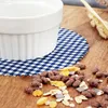 A close-up of a white ramekin dish on a blue and white checkered cloth, surrounded by scattered cereal pieces and chocolate squares on a wooden surface. Produktbild-Vorschau