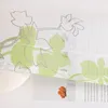 A close-up of a sheer curtain with a green and gray floral pattern, partially covering a white ribbed object and a piece of paper with text and an orange dried flower. Produktbild-Vorschau