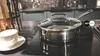 A close-up shot of a stainless steel pot with a glass lid on a black induction stovetop, with a white teacup and saucer to the left. Produktbild-Vorschau