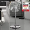 A chrome stand fan is shown in a modern living room setting, with a sofa and a floor lamp in the background. A promotional badge is visible on the right. Podgląd zdjęcia produktu