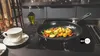 A frying pan with vegetables cooking on a stovetop, with a cup and saucer to the side. Produktbild-Vorschau