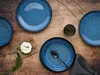 A top-down view of a table setting with blue plates, a small cutting board with a dollop of cream cheese and dill, a glass of dark liquid, and a dark napkin with a spoon. Produktbild-Vorschau