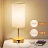 A bedside lamp with USB charging ports is shown on a nightstand next to a smartphone and a small charging brick. Produktbild-Vorschau