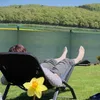 A person relaxing in a lounge chair outdoors, with a fence and trees in the background. Podgląd zdjęcia produktu