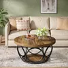 A round coffee table with a wooden top and black metal base is placed in a living room setting in front of a sofa. Anteprima dell’immagine prodotto