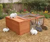 A wooden rabbit hutch with several rabbits in a garden setting with straw and flowers. Produktbild-Vorschau