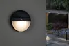 A split image showing a close-up of a black and white solar LED outdoor wall light on the left, and a nighttime view of a modern garage with landscaping and pathway lighting on the right. Náhled obrázku produktu