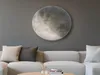 A large circular light fixture resembling the moon is mounted on a gray wall above a white sectional sofa. Two pillows, one beige and one dark gray, are on the sofa. A small vase with flowers and a book are on a coffee table in front of the sofa. Náhled obrázku produktu