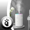 A white humidifier with a colorful LED light is shown emitting mist, with white roses in the background. A black and white graphic of a hand making an 'okay' sign is in the foreground. Produktbild-Vorschau