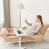 A pregnant woman is relaxing on a chaise lounge, using a laptop and her phone. A floor lamp and curtains are visible in the background. Produktbild-Vorschau