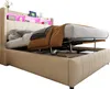 A beige upholstered bed with its storage mechanism partially open, revealing storage space underneath. The headboard features LED lighting and shelves. Náhled obrázku produktu
