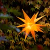 A glowing orange star lantern hangs in front of a wall covered in autumn leaves. Produktbild-Vorschau