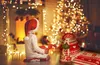A child in a Santa hat sits on the floor in front of a fireplace and a Christmas tree, holding a gift. Anteprima dell’immagine prodotto