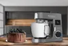 A kitchen mixer and a chocolate cake are displayed on a wooden table in a kitchen setting. Produktbild-Vorschau