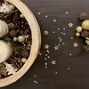 A close-up view of a Christmas-themed centerpiece with candles, pinecones, and gold star confetti scattered on a dark wooden surface. Produktbild-Vorschau