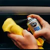 A person is spraying a cleaning product onto a yellow microfiber cloth, which is held in front of a dark surface, likely a car interior. Podgląd zdjęcia produktu
