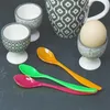 A collection of egg cups, a mug, an egg, and colorful spoons arranged on a dark gray surface. Produktbild-Vorschau