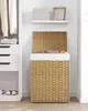 A woven laundry basket with its lid open, placed next to a white closet with clothes and drawers. Náhľad obrázku produktu