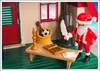 A Playmobil Santa Claus figure in a festive setting, holding a quill and standing next to a desk with an open book. Produktbild-Vorschau