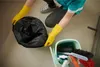 A person wearing yellow gloves is holding a black trash bag next to a cleaning bucket filled with cleaning supplies. Produktbild-Vorschau