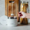 A hand is scooping food from a white insulated food container, with a jar of spaghetti and a wooden cutting board in the background. Voir les images du produit