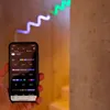 A hand holding a smartphone displaying a lighting control app, with a colorful LED light strip installed on a wall in the background. Produktbild-Vorschau