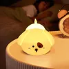 A cute dog-shaped LED night light is placed on a bedside table next to a sleeping person and an alarm clock. Produktbild-Vorschau