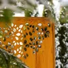 A rusty metal garden screen with a floral cut-out pattern is partially obscured by green branches and snow-covered foliage in the background. Produktbild-Vorschau