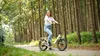 A woman is riding an electric bicycle on a paved path through a forest. Produktbild-Vorschau