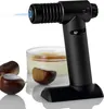 A black culinary torch is shown in use, with a flame directed towards a crème brûlée in a ramekin. Several chestnuts are also visible in the foreground. Produktbild-Vorschau