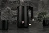 A black electric kettle and a cup are placed on a dark countertop with a stone-like wall in the background and a small bonsai tree to the right. Náhľad obrázku produktu