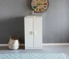 A white cabinet with two doors stands against a gray wall, next to a woven basket. A clock hangs on the wall above the cabinet. Produktbild-Vorschau