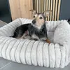 A small dog is lying in a plush, white dog bed. The background shows a modern interior with wooden paneling. Produktbild-Vorschau