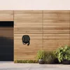 A black electric vehicle charging station mounted on a wooden wall, with plants in pots in the foreground. Anteprima dell’immagine prodotto