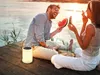 A couple is sitting on a wooden dock by the water at sunset, eating watermelon. A solar lantern is placed on the dock next to them. Produktbild-Vorschau