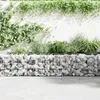A gabion planter filled with rocks and plants, set against a white wall with climbing vines. Produktbild-Vorschau
