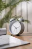 A blue analog alarm clock with gold accents sits on a wooden table next to a rolled-up document. Anteprima dell’immagine prodotto