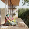 A colorful bean bag chair sits on a wooden deck next to a small woven stool, with plants and a stone wall in the background. Voir les images du produit