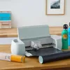 A Cricut Explore 3 cutting machine is shown on a wooden table, along with rolls of vinyl and water bottles. Produktbild-Vorschau
