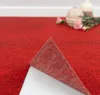 A close-up of a red carpet tile being peeled back, revealing its adhesive backing. The background shows a white baseboard and some decorative vases and flowers. Produktbild-Vorschau