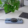 A blue iPhone is charging on a white desk with a wireless charger attached. A keyboard and a plant are visible in the background. Anteprima dell’immagine prodotto