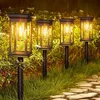 Four solar garden lights are illuminated at dusk, lining a stone pathway through a garden. Produktbild-Vorschau