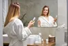 A woman in a bathrobe is holding a bottle of skincare product in front of a bathroom mirror. Produktbild-Vorschau